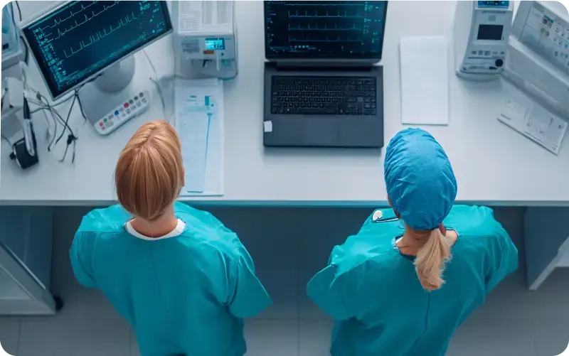 An image showing two healthcare professionals in light blue uniforms looking at a laptop screen.