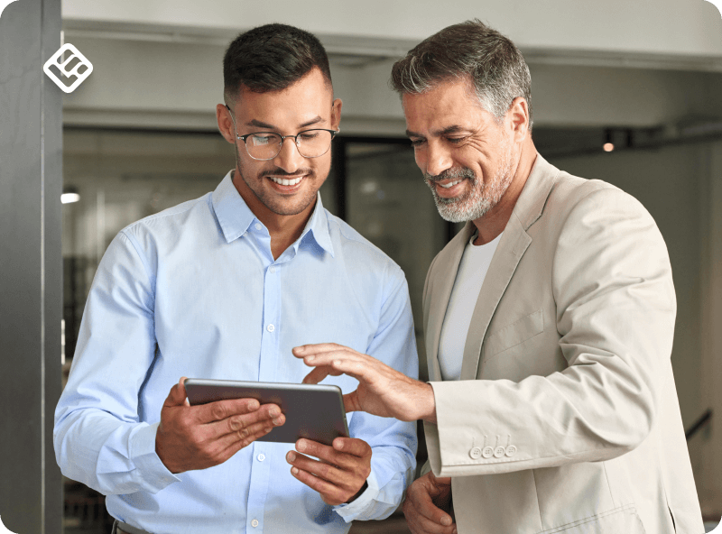  A screenshot showing two men smiling while looking at something on and holding a tablet.